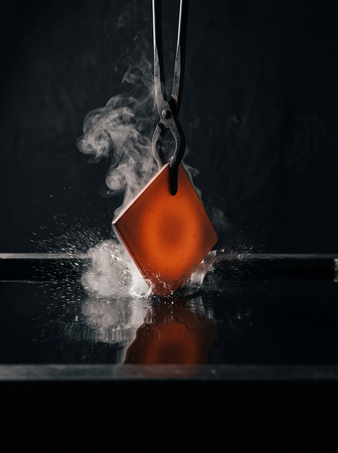 A furnace-hot glass-ceramic plate held in steel tongs entering a black water bath, steam and spray frozen at the moment of contact